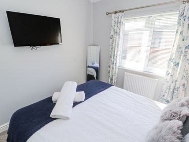 A bedroom with a wall mounted television two white towels on a bed with a blue and white blanket a window with floral curtains and a full length mirror at Duckling Lodge in South Lakeland Leisure Village