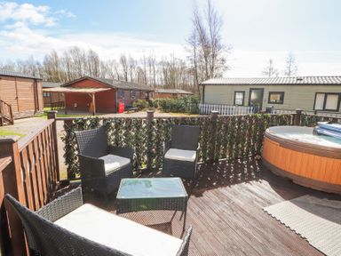 An outdoor deck with wicker chairs a glass table and a hot tub surrounded by wooden lodges at South Lakeland Leisure Village