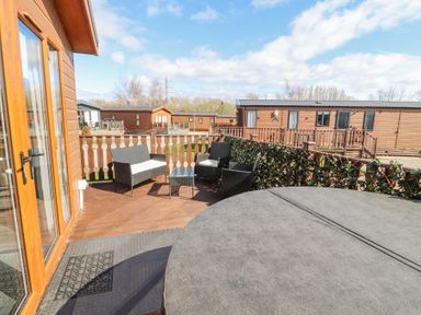 An outdoor deck with black wicker chairs a glass table and a hot tub next to wooden lodges at Duckling Lodge in South Lakeland Leisure Village