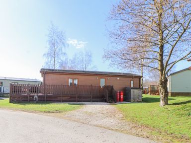 A wooden lodge with a deck and steps surrounded by grass and trees at Duckling Lodge in South Lakeland Leisure Village