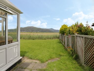 A garden with grass and fenced plants next to a conservatory with hills in the background at Hafod-y-Gors in Fairbourne