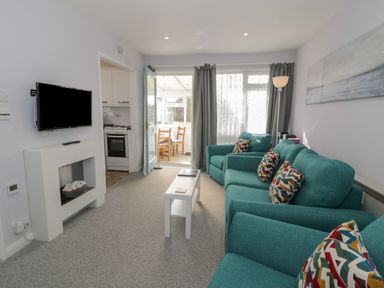A living room with teal sofas and multicolored cushions with a white coffee table and a wall-mounted TV at Hafod-y-Gors in Fairbourne