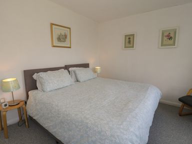 A bedroom with a large bed with light patterned bedding two bedside tables with lamps and framed pictures on walls at Hafod-y-Gors in Fairbourne