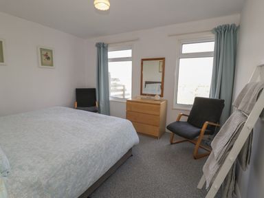 A bedroom with two black chairs a wooden chest of drawers a mirror two windows and a bed with a light blue bedspread at Hafod-y-Gors in Fairbourne