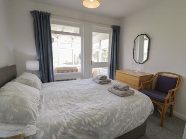 A bedroom with a bed folded towels on top a wooden chest of drawers a wicker chair and a small lamp at Hafod-y-Gors in Fairbourne