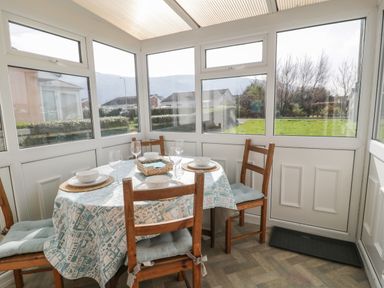 A dining area with a round table set four with dishes and glasses and wooden chairs in a sunroom at Hafod-y-Gors in Fairbourne