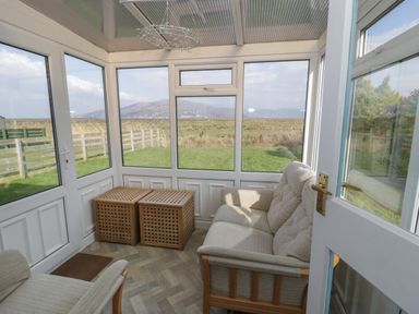 A sunroom with two armchairs and two wooden side tables overlooking a grassy yard at Hafod-y-Gors in Fairbourne