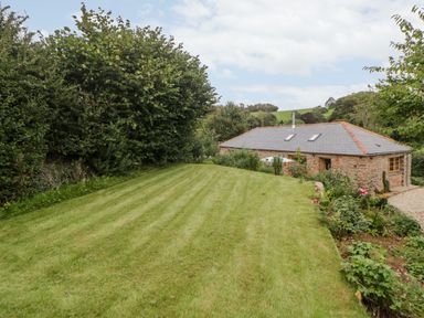A grassy lawn with trees and bushes surrounding a stone house with a slate roof at The Old Haybarn in Ipplepen
