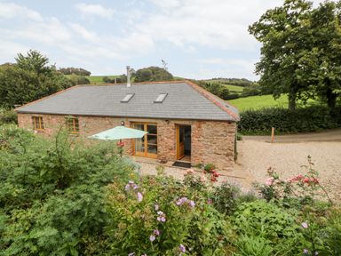 A stone cottage with a grey slate roof surrounded by green bushes and colorful flowers at The Old Haybarn in Ipplepen