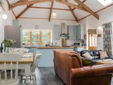 An open plan kitchen and living area with wooden beams dining table and chairs a brown leather sofa a black sofa and kitchen cabinets at The Old Haybarn in Ipplepen