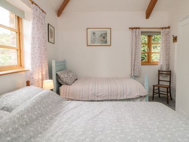 A bedroom with two beds a lamp a wooden chair and windows with curtains at The Old Haybarn in Ipplepen
