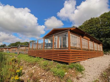 A row of wooden cabins with glass and wood railings on raised decks surrounded by grass and trees at Mereside South lakeland Leisure Village near Carnforth