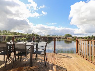An outdoor wooden deck with a dining table and chairs overlooking a lake and cabins at Mereside in South lakeland Leisure Village near Carnforth