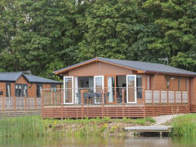 Wooden cabins with deck and outdoor furniture near a pond surrounded by trees at Mereside South lakeland Leisure Village near Carnforth