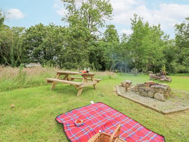 An outdoor picnic scene with a wooden picnic table wine glasses a fire pit and a red checkered blanket on grass at The Farmhouse in Newport Pembrokeshire