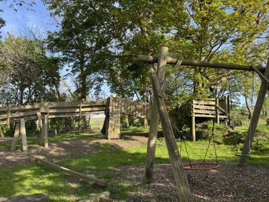 A wooden swing set and playground equipment with trees and grass at The Farmhouse in Newport Pembrokeshire