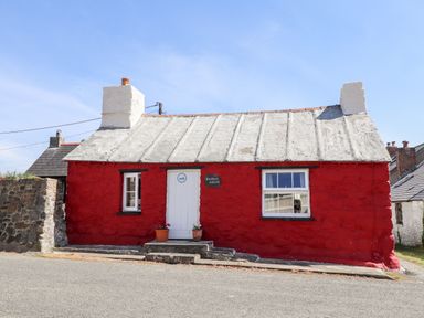 A small red stone cottage with a white door and two windows on a street at Bwthyn Alarch in Trefin St Davids Pembrokeshire