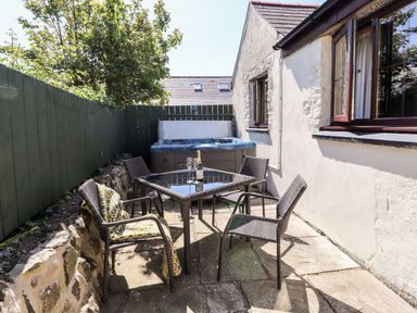 An outdoor patio with four chairs around a glass table and a hot tub against a white stone building at Bwthyn Alarch in Trefin St Davids Pembrokeshire