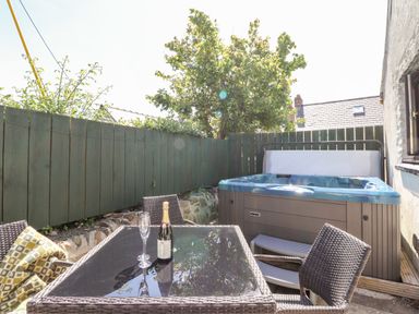 An outdoor patio with a glass table and wicker chairs a bottle of wine two glasses a green wooden fence and a hot tub at Bwthyn Alarch in Trefin St Davids Pembrokeshire
