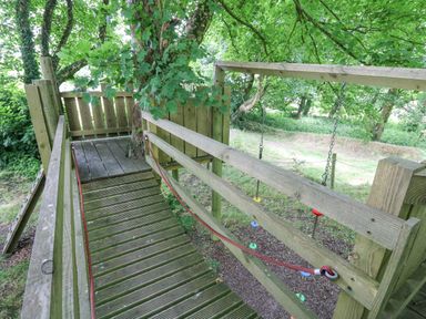 A wooden treehouse platform with a slanted wooden bridge and rope on the side surrounded by trees at The Granary in Newport Pembrokeshire