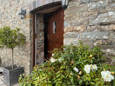 An open wooden door in a stone wall with plants and white flowers outside at The Granary in Newport Pembrokeshire