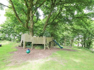 An outdoor playground with a wooden play structure around a tree a slide a swing and wood chips on the ground at Stable Cottage in Newport Pembrokeshire