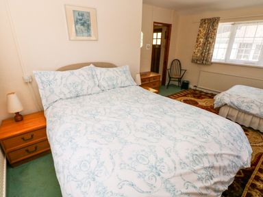 A bedroom with two beds a wooden nightstand with a lamp a wooden chair a framed picture on the wall and a window at Otter Cottage in Dinas Cross