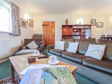 A living room with a brown sofa with white cushions a brown armchair a wooden door a wooden bookshelf a wooden coffee table with cups and magazines at Otter Cottage in Dinas Cross