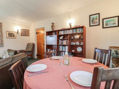 A dining table set with plates and glasses in a room with a wooden bookcase and armchairs at Otter Cottage in Dinas Cross