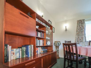 A dining room with wooden chairs a table with a pink tablecloth a wooden bookshelf with books and china at Otter Cottage in Dinas Cross
