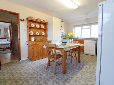 A kitchen with wooden table and chairs a wooden cabinet with plates on the wall a dishwasher and washing machine near a window at Otter Cottage in Dinas Cross