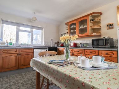 A kitchen with wooden cabinets a table with a floral tablecloth cups on a tray and a vase of flowers at Otter Cottage in Dinas Cross