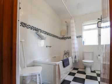 A bathroom with a bathtub shower toilet white chair and checkered floor at Otter Cottage in Dinas Cross