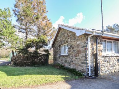 Exterior view of a stone house with windows and a metal gate next to a lawn and trees at Otter Cottage in Dinas Cross