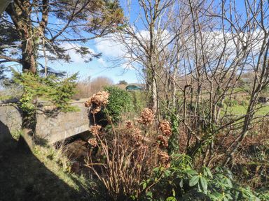 A garden with trees shrubs and dried flowers near a stone bridge over a stream at Otter Cottage in Dinas Cross