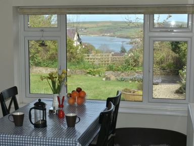 A dining area with a table covered by a checkered cloth and chairs by a large window with a view of a garden and water at Gellifor in St Dogmaels Pembrokeshire