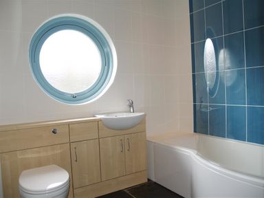 A bathroom with a round window a toilet a sink with wooden cabinets and a bathtub with blue tiled wall at Seabrook Broad Haven Pembrokeshire