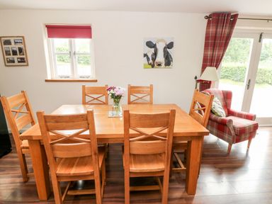 A dining room with a wooden table and six chairs a vase with flowers a red plaid armchair and a window with red curtains at Gwyddno Lodge in Narberth Pembrokeshire