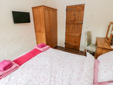 A bedroom with a bed covered in pink floral bedding a wooden wardrobe a wooden door a wall-mounted TV and a desk with a chair at Gwyddno Lodge in Narberth Pembrokeshire