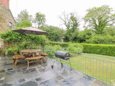 A patio with a wooden picnic table and umbrella and a barbecue grill overlooking a garden at Greenbanks in Dinas Cross Pembrokeshire
