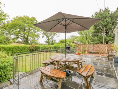 A wooden picnic table with umbrella on a wet paved patio overlooking a garden with trees and hedges at Greenbanks in Dinas Cross Pembrokeshire