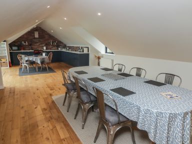 A dining area with a long table covered with a blue and white tablecloth and metal chairs and a kitchen with a smaller round table in an attic space at The Willows in Newport Pembrokeshire