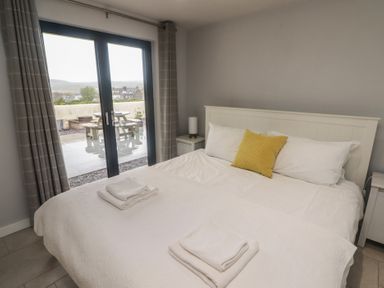 A bedroom with a bed with white bedding and a yellow cushion and two folded towels on the bed by a glass door with curtains at The Willows in Newport Pembrokeshire
