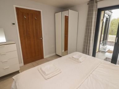 A bedroom with a bed with folded towels on it a white dresser a wooden door a white wardrobe with a mirror and sliding glass doors with curtains at The Willows in Newport Pembrokeshire