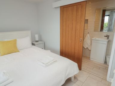 A bedroom with a white bed with folded towels and a yellow pillow next to a wooden sliding door leading to a small bathroom with a sink at The Willows in Newport Pembrokeshire