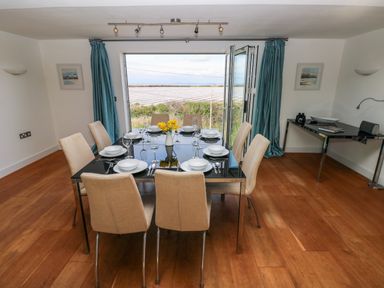 A dining room with a set table and beige chairs next to a large open window with blue curtains at Gwelfor Marloes in Marloes Pembrokeshire