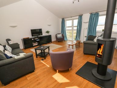 A living room with sofas chairs a television on a stand a wood stove and two tables with blue curtains at Gwelfor Marloes in Marloes Pembrokeshire