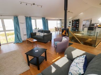 An open living room with sofas and armchair around a central fireplace with a glass staircase and kitchen in the background at Gwelfor Marloes in Marloes Pembrokeshire
