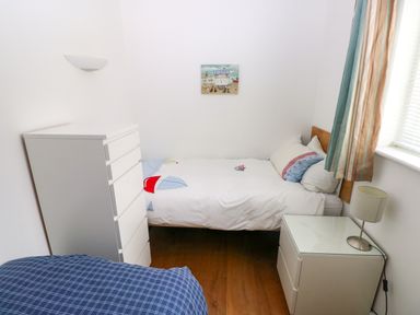 A small bedroom with two beds a white chest of drawers a bedside table with a lamp and a window with curtains at Gwelfor Marloes in Marloes Pembrokeshire