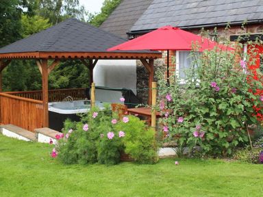A garden with pink flowers a wooden gazebo with a hot tub a red umbrella and a stone house at Pond Cottage in New Quay Ceredigion